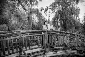 Black and white photo of a couple standing on a rustic wooden bridge surrounded by lush trees.