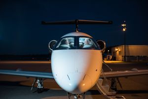 Front view of a white private jet with two people visible through cockpit windows at night.