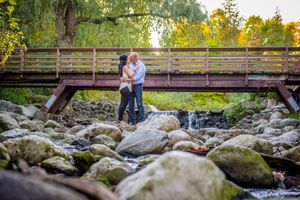 Couple sharing a kiss on a wooden bridge over a rocky waterfall surrounded by autumn foliage.