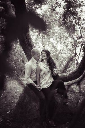 Black and white photo of a romantic couple sitting together on a large tree root in a forest.
