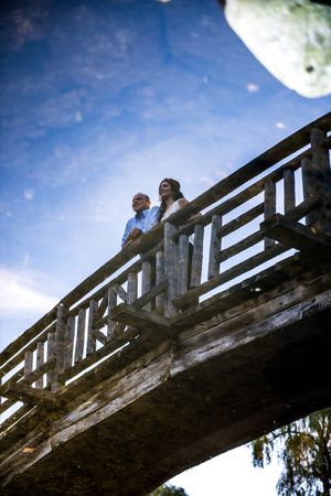 Low-angle reflection shot of a couple standing on a wooden bridge against a bright blue sky.