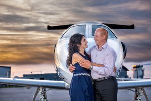 Couple embracing and smiling in front of a private jet on an airport tarmac at colorful sunset.