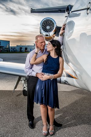 Couple smiling together beside a private jet on a tarmac at sunset, woman in blue dress.