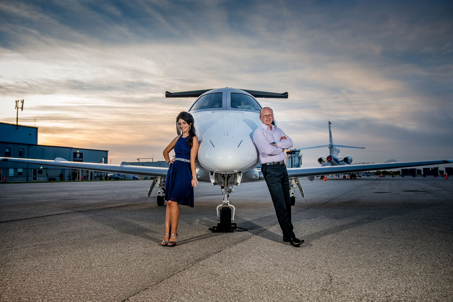 Couple standing apart in front of a private jet on an airport tarmac at dramatic sunset.