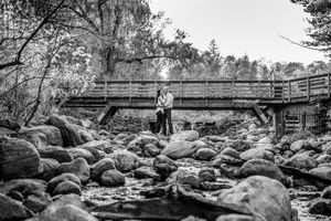 Black and white photo of a couple embracing on a rocky riverbed beneath a wooden bridge.