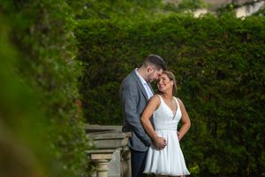 Couple sharing a tender moment near stone balustrade surrounded by lush green hedges in a garden setting.