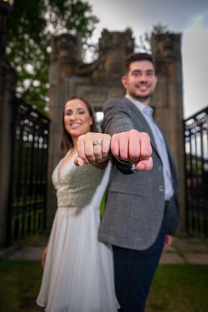 Smiling engaged couple showing off their rings by extending fists toward camera in front of ornate iron gates.