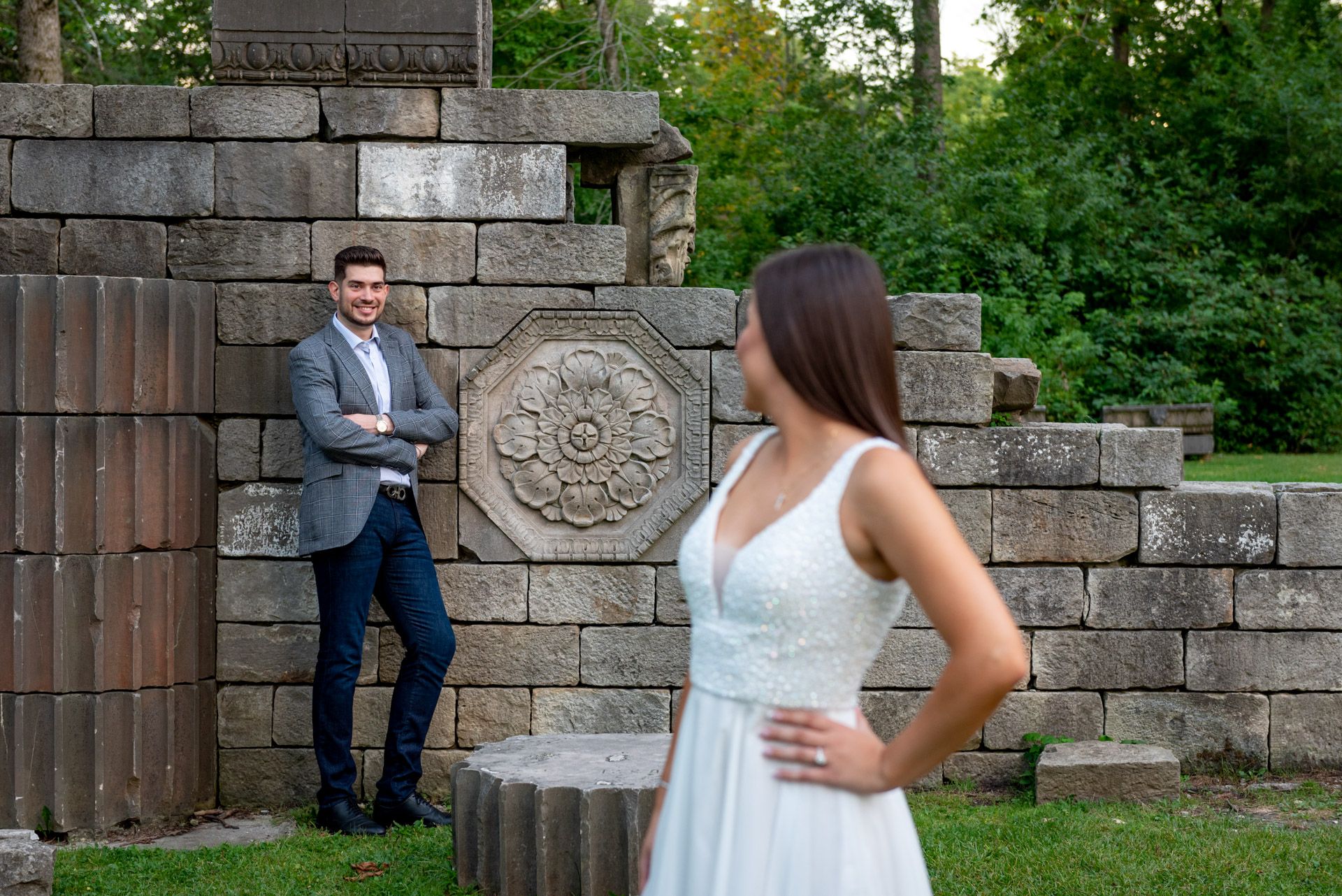 Man in gray blazer leaning against ornate stone wall as woman in white dress stands facing him in a park.