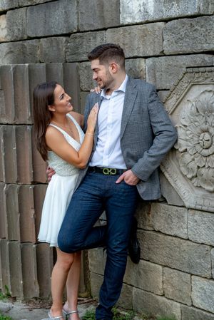 Engaged couple smiling at each other beside an ornate stone wall, woman in white dress touching mans gray blazer.