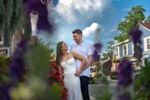 Couple in love standing close together amid vibrant purple and red flowers with a bright cloudy sky above.
