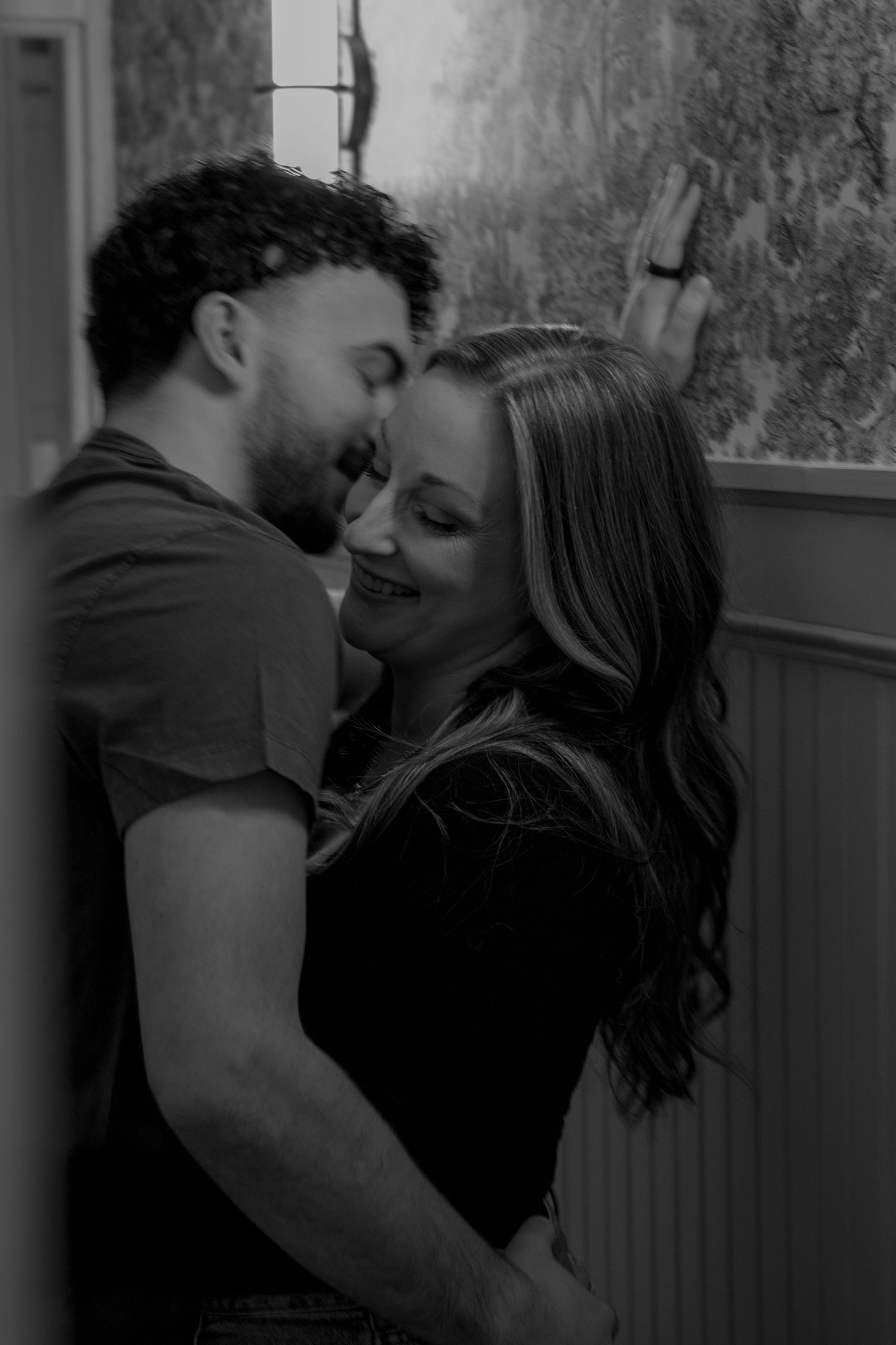 Romantic black and white photo of a couple sharing an intimate moment near a window indoors.