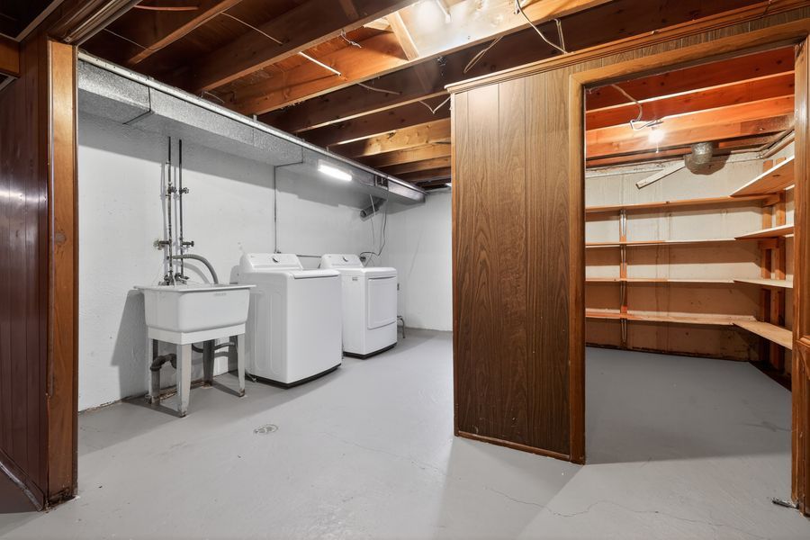 Unfinished basement laundry room with utility sink, washer, dryer, and adjacent wood-paneled storage room with shelving.