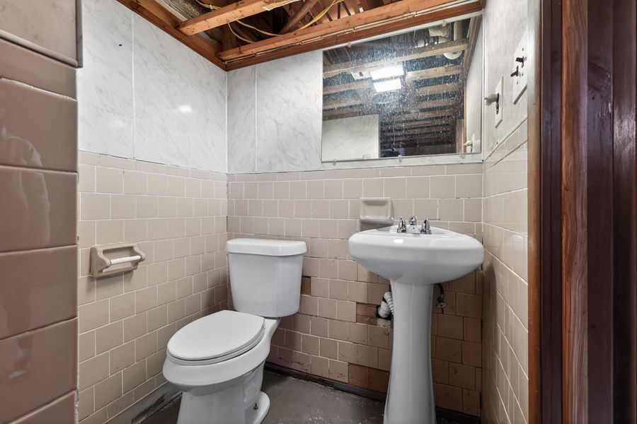 Unfinished basement bathroom with white toilet, pedestal sink, tile walls, and exposed ceiling joists.