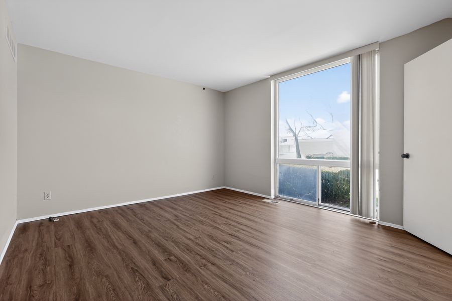 Bright empty room with dark hardwood floors, neutral walls, and large floor-to-ceiling window with outdoor view.