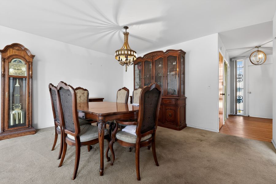 Elegant dining room with wooden table, cane-back chairs, china cabinet, grandfather clock, and chandelier on carpeted floor.