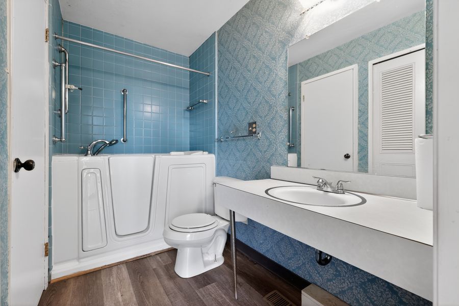 Modern bathroom with blue tile walls, walk-in tub, white toilet, and single vanity sink on dark hardwood floors.