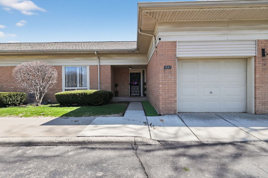 Brick townhouse with attached garage, covered entryway, manicured shrubs, and concrete driveway under a partly cloudy sky.