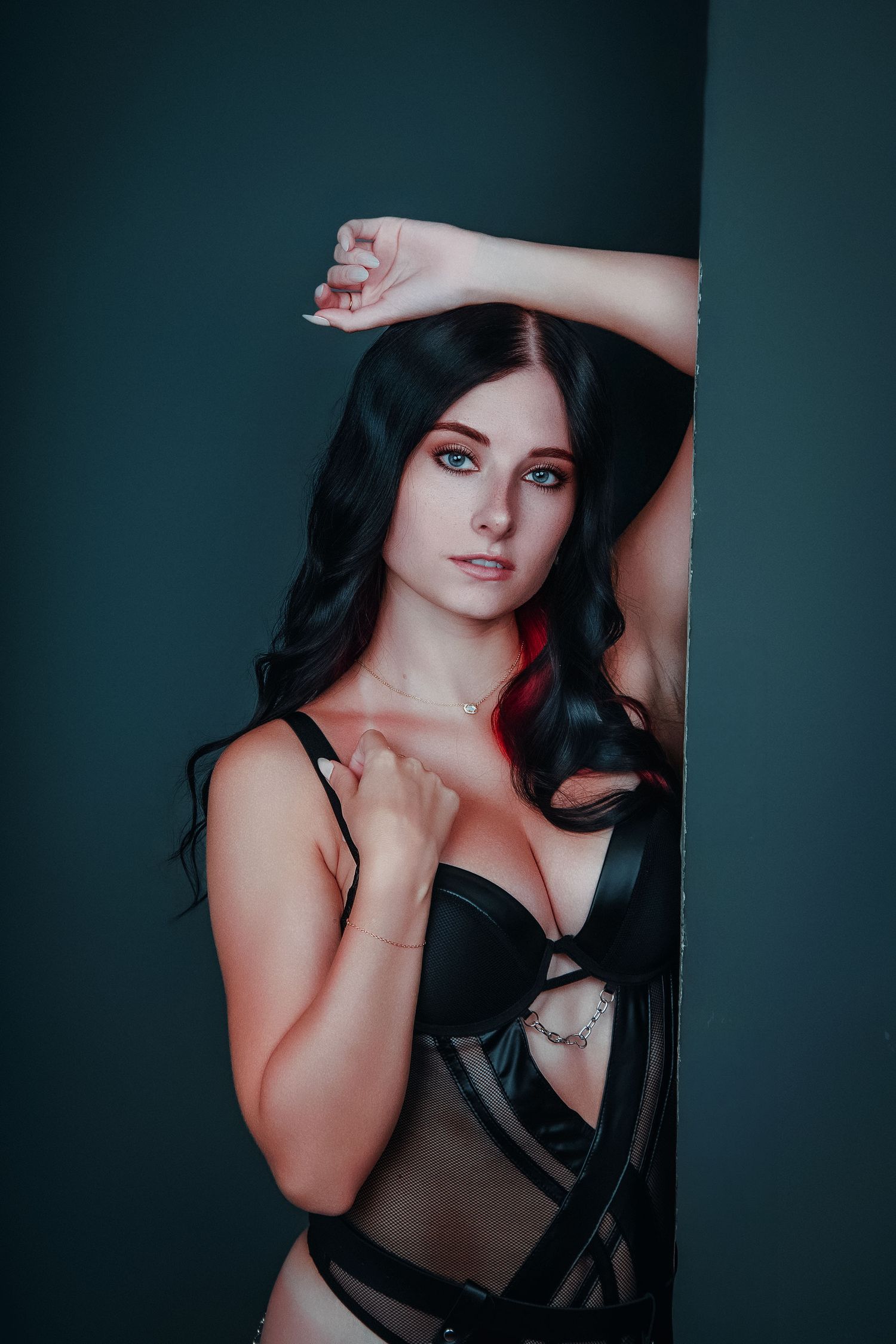 Young woman with dark hair posing in black lingerie against a teal wall in a studio boudoir photo.