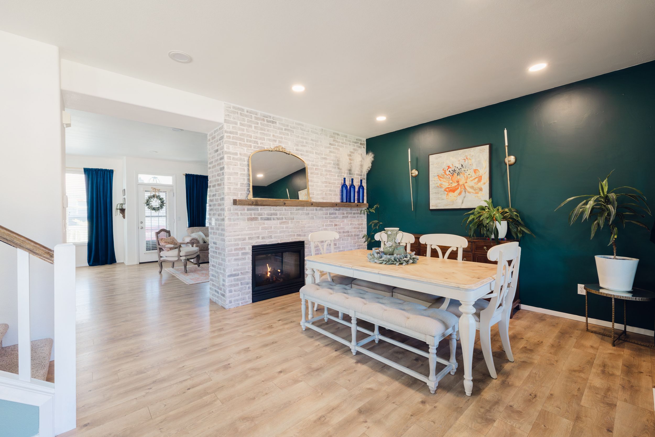 Open-concept dining room with teal accent wall, white brick fireplace, farmhouse table, and light hardwood floors.