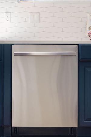 Stainless steel dishwasher with bar handle beneath white hexagon tile backsplash and navy blue cabinets.