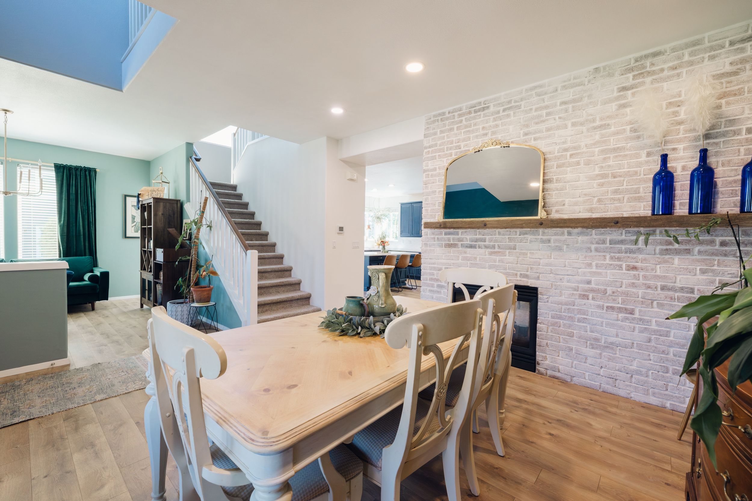 Bright dining room with wooden table, white chairs, brick fireplace, and staircase leading to upper level.