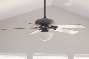Modern ceiling fan with five blades and glass light fixture mounted on vaulted white ceiling in a bright room.