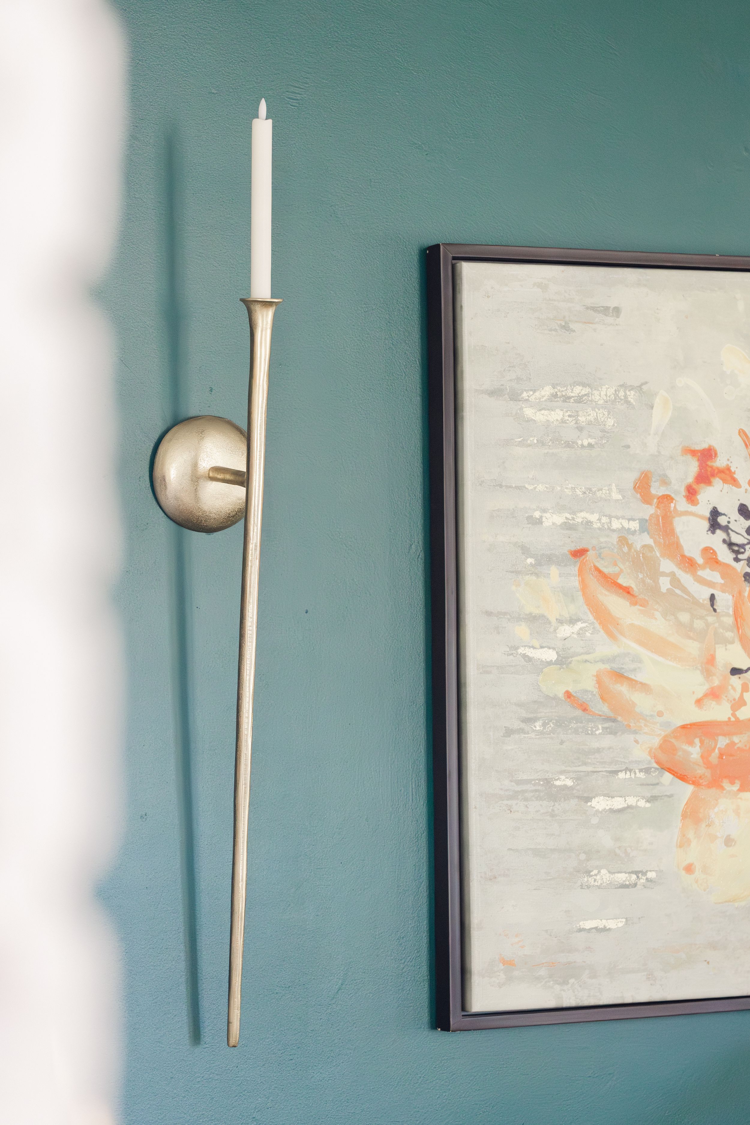 Gold taper candle wall sconce beside abstract floral artwork on teal wall.