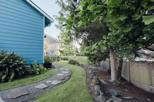 Curved stone garden path along blue house with lush green landscaping and wood fence.