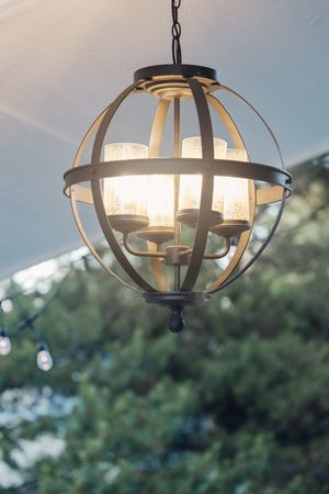 Elegant spherical bronze outdoor chandelier with candle-style lights hanging against a backdrop of green trees.