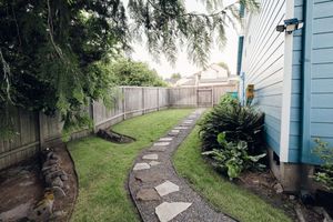 Narrow backyard pathway with stepping stones, lush green lawn, wooden fence, and tropical plants beside blue house exterior.