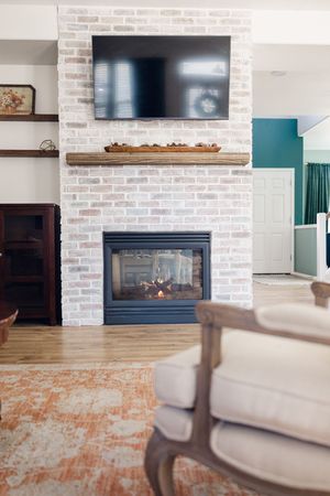 Whitewashed brick fireplace with wood mantel, mounted TV, and glowing fire in a cozy living room with hardwood floors.