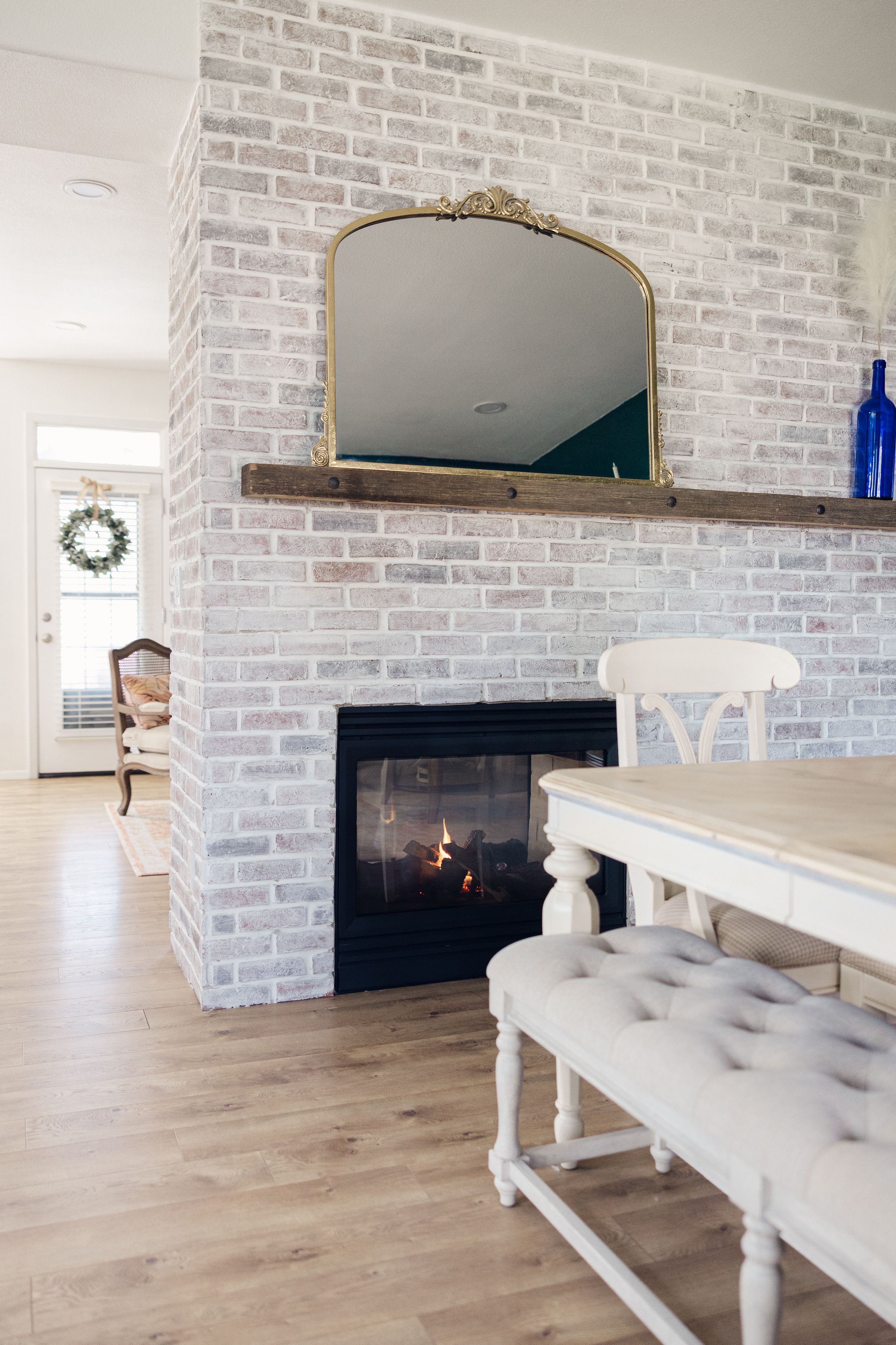 Whitewashed brick fireplace with wood mantel, gold mirror, and blue vase in a bright dining room with hardwood floors.