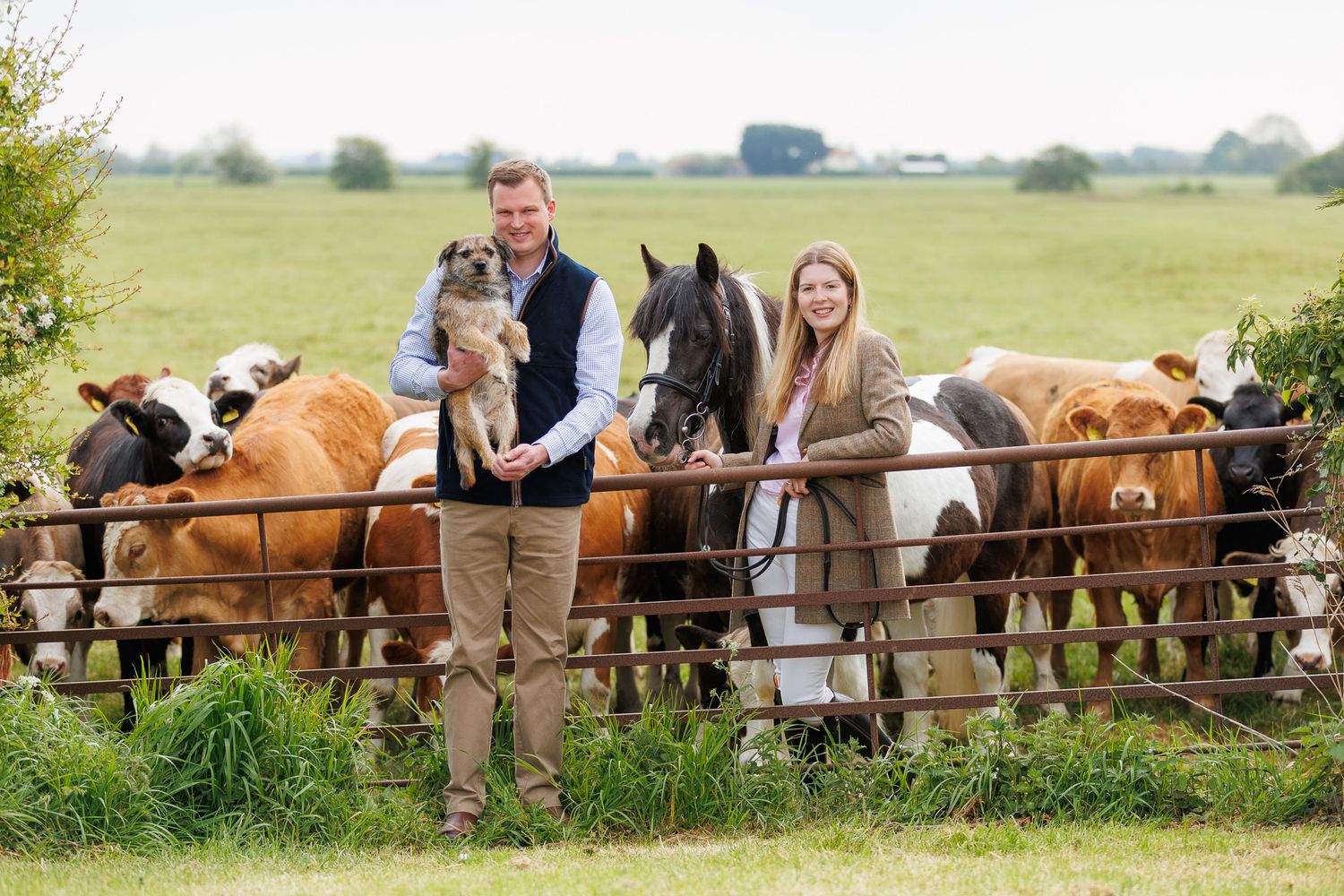 A man and woman smile at a farm gate surrounded by cattle and a horse in a lush green field.