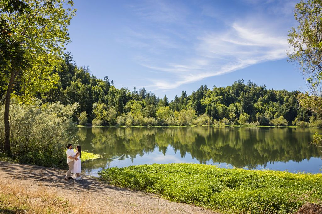Two people stand by a serene lake surrounded by lush green trees on a sunny day.