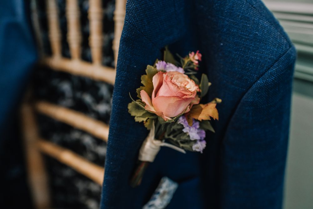 A pink rose boutonniere with purple accents pinned to a navy blue suit jacket at a wedding.