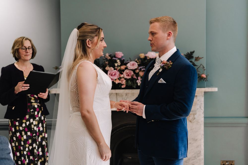 Bride and groom exchange rings at altar during indoor wedding ceremony with officiant and floral arrangement in background.