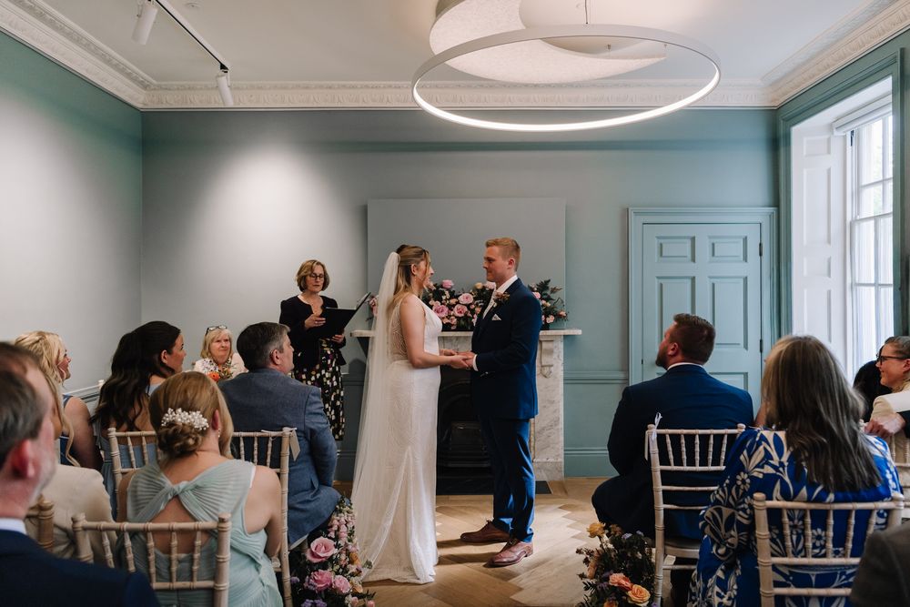 Bride and groom exchange vows during indoor wedding ceremony in elegant blue-walled venue with seated guests.