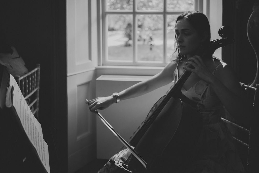 Black and white photo of a woman playing cello near a window, captured in an elegant indoor setting.