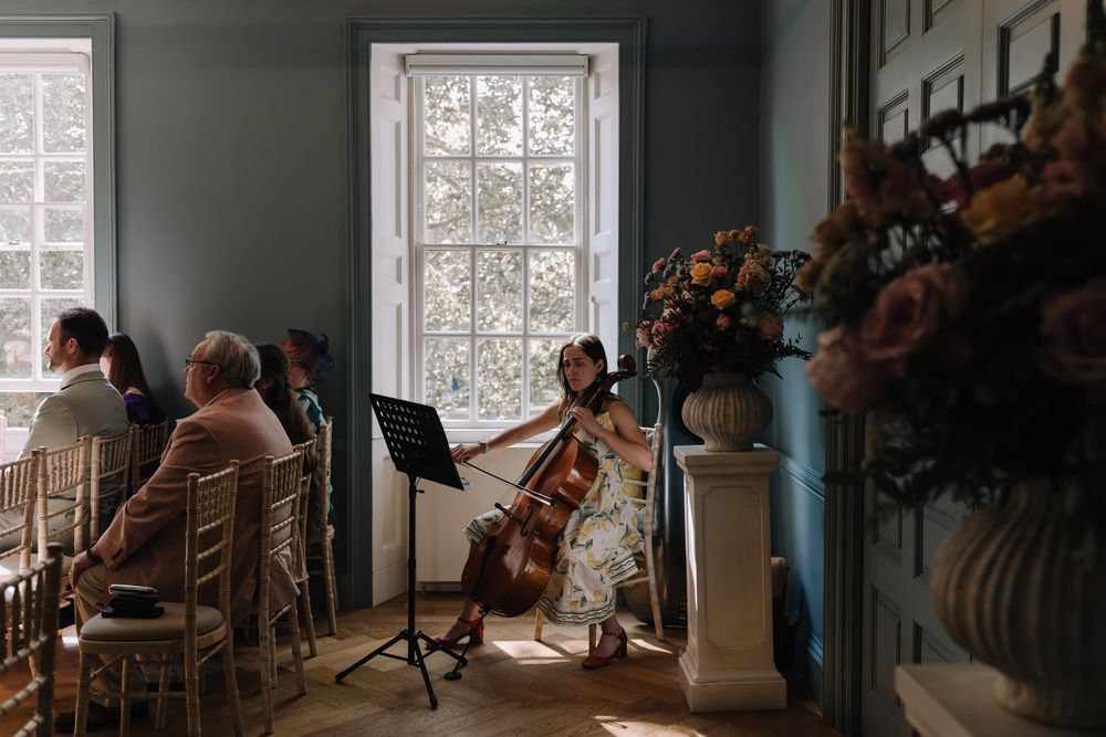 A cellist performs at an elegant wedding ceremony in a blue-walled room with guests seated nearby.