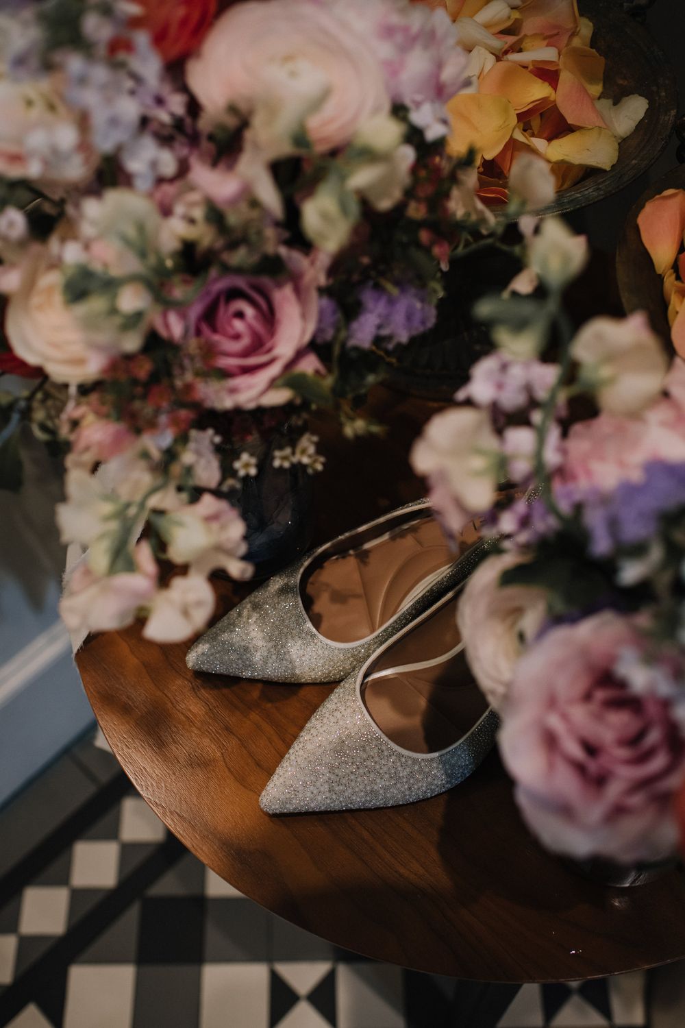 Glittery bridal heels displayed on a wooden table surrounded by colorful floral arrangements with pink and purple blooms.