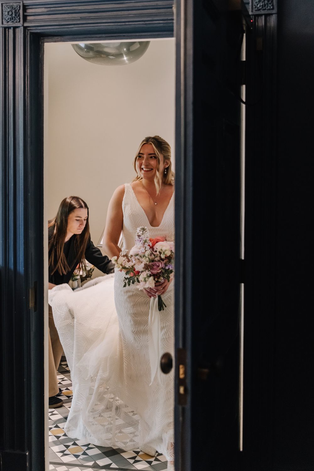Bride in white gown holds pink bouquet while assistant adjusts dress in elegant doorway.