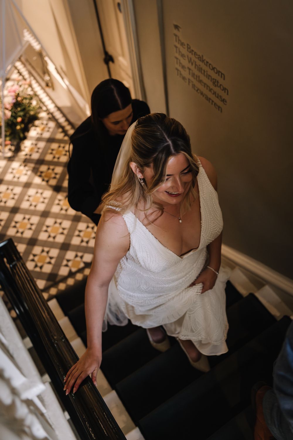 Bride in white gown climbs staircase with assistant helping carry her dress train.