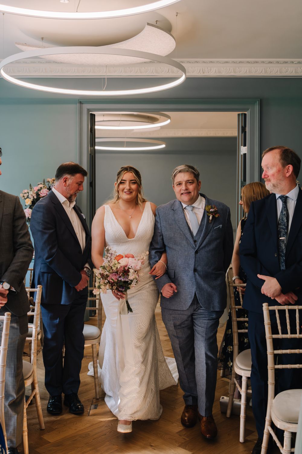 Bride walks down the aisle with her father in a elegant wedding venue with teal walls.