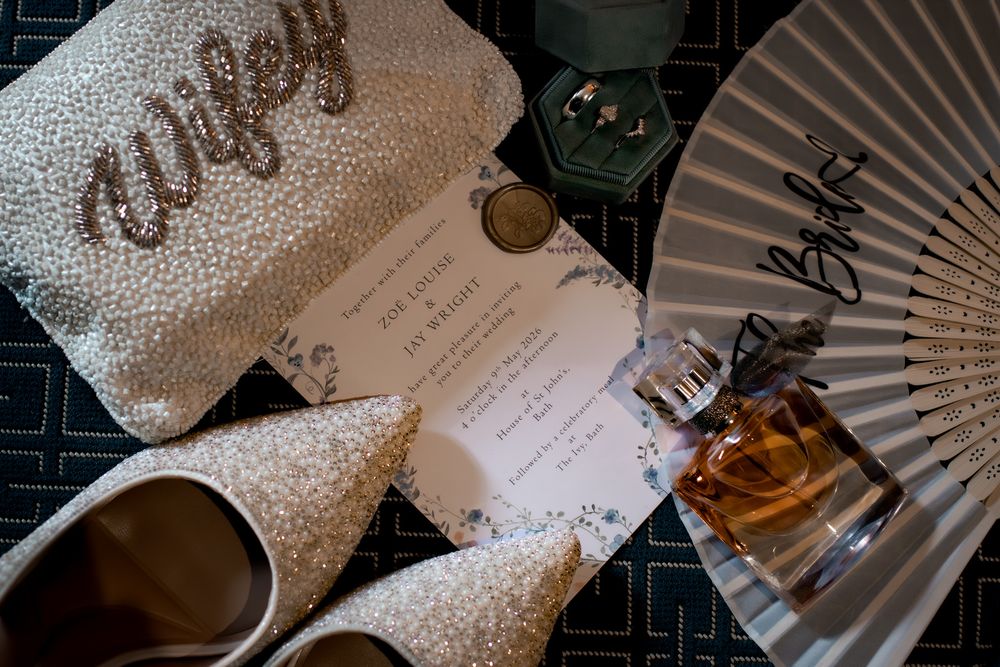 Bridal flat lay featuring glittery heels, wifey clutch, wedding invitation, perfume bottle, and fan on dark surface.