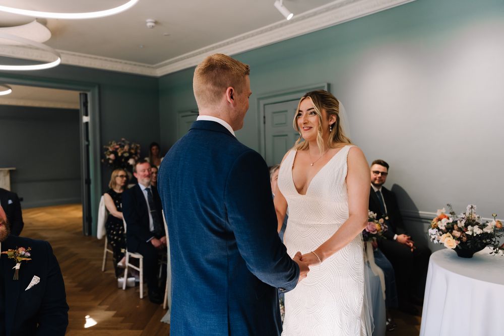 Bride and groom holding hands during intimate wedding ceremony in elegant teal-walled venue with guests seated behind.