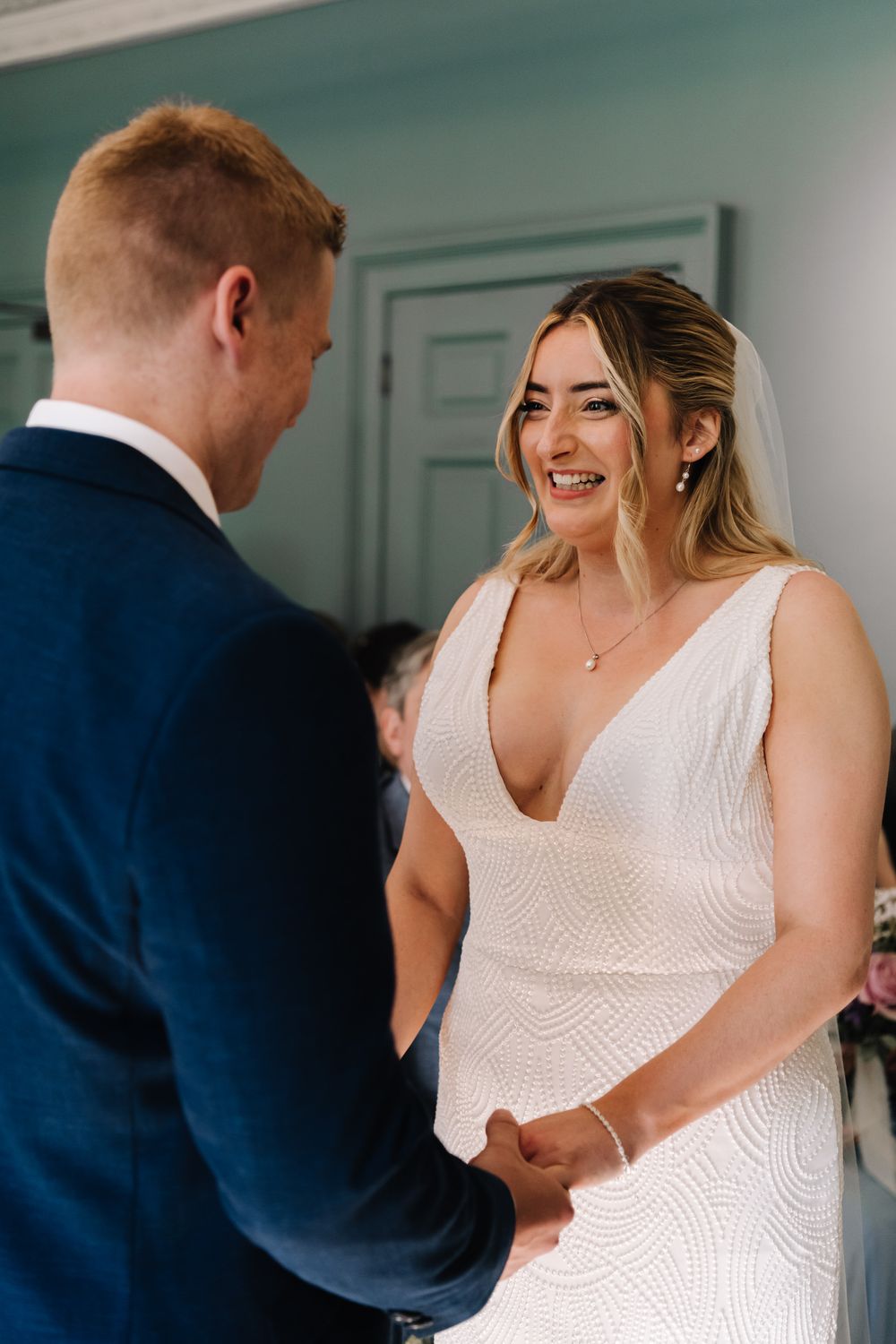 Bride in white gown smiling joyfully at groom in navy suit while holding hands during wedding ceremony.
