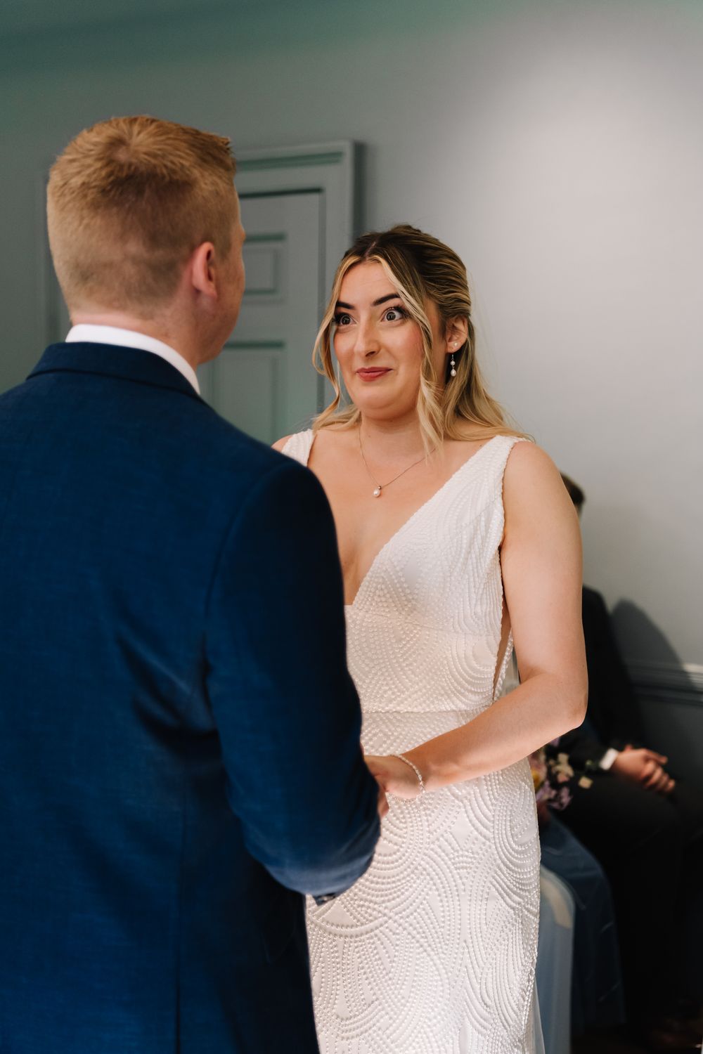 Bride in a beaded gown exchanges vows with groom in navy suit at wedding ceremony.