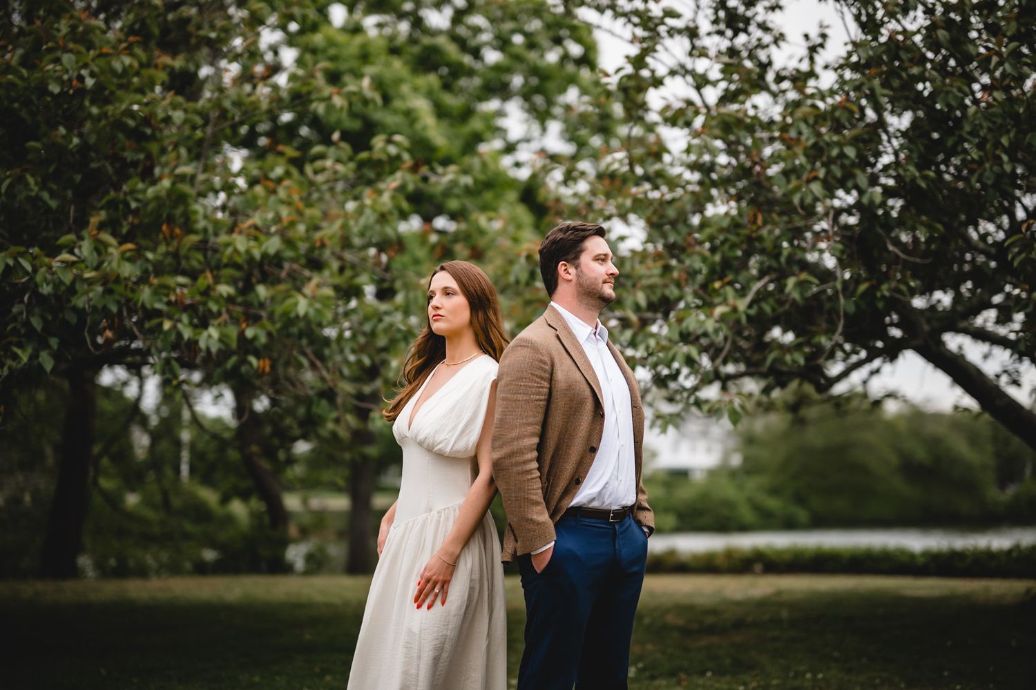 A couple in wedding attire stand back-to-back in a lush green park, the woman in white dress, man in brown jacket.