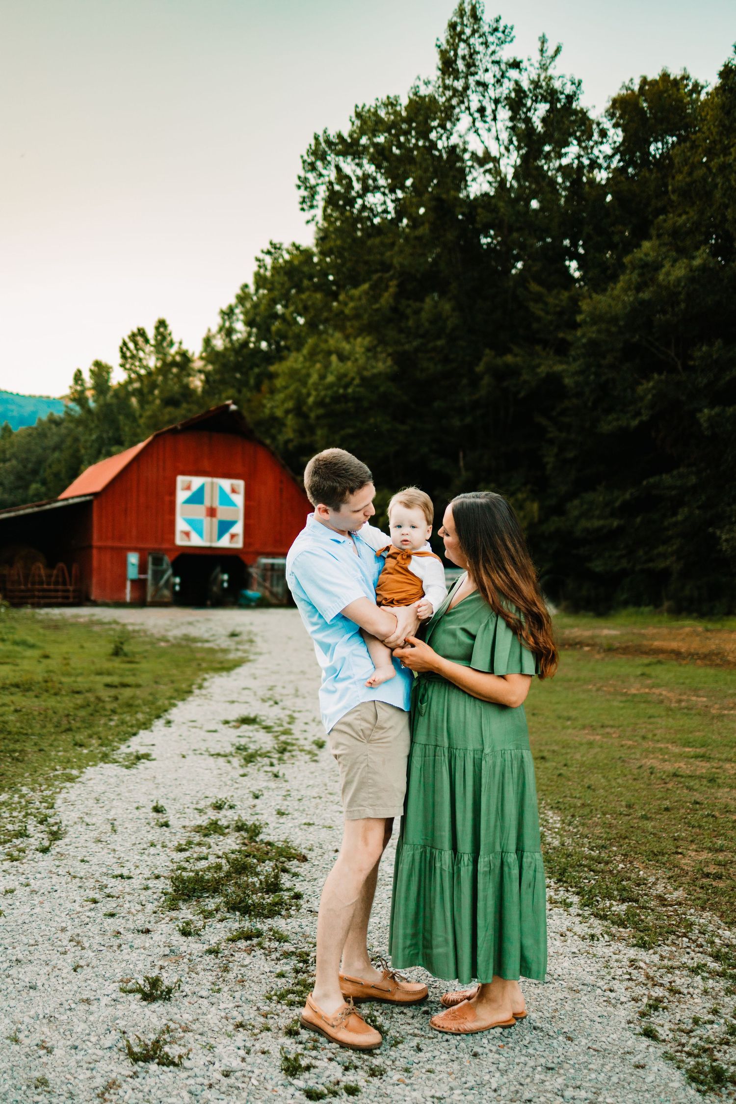 A young family with a baby poses on a gravel path before a red barn and lush green trees at golden hour.