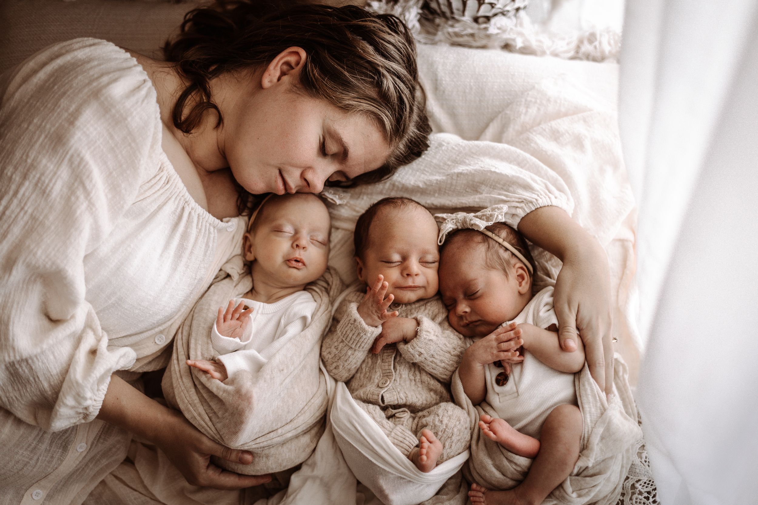 Mother tenderly kisses sleeping newborn triplets wrapped in white swaddles on a soft bed.